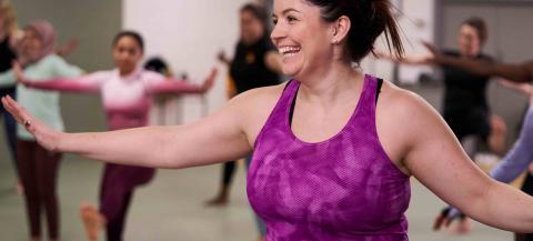 A woman is taking part in a movement class with other women and girls, She is wearing a purple top and she is smiling. 